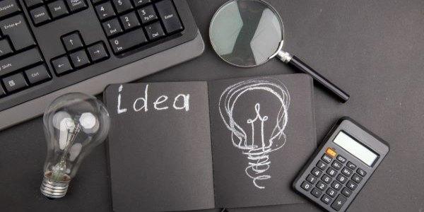 Top view idea written on black notepad keyboard lupa binder clip light bulb calculator on dark