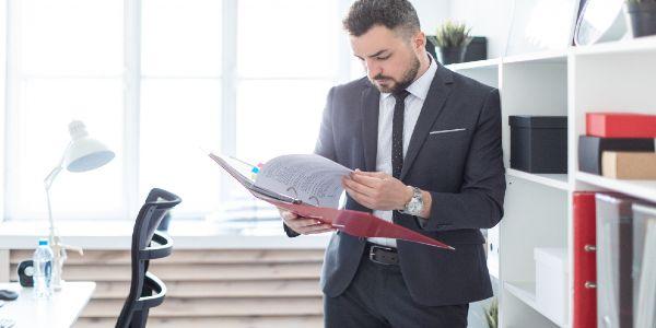 The man is standing near the rack in the office and scrolls through the folder with the documents