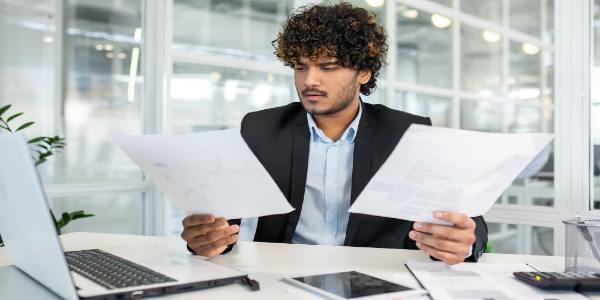 Focused young businessman with curly hair wearing a suit examining multiple documents and reports