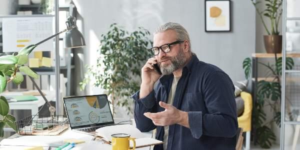 Mature businessman in eyeglasses sitting at his workplace and talking on mobile phone at office