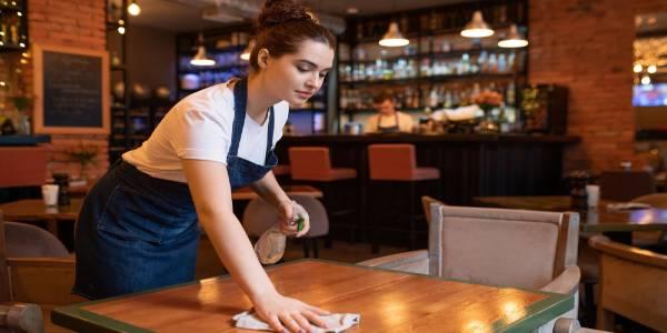 Young waitress in apron cleaning wooden table using detergent and duster in restaurant