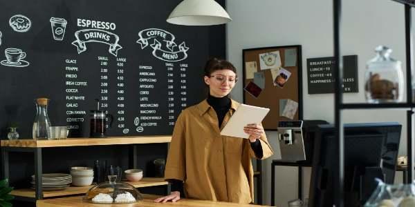 Young smiling woman looking at tablet screen while standing by counter desk with cafe menu on wall behind
